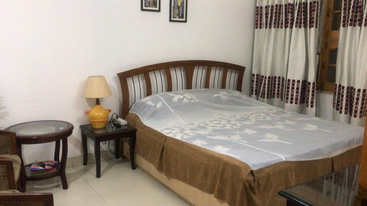 A cozy bedroom features a wooden bed with a light bedspread and matching throw. A bedside table with a lamp is positioned beside the bed. Window curtains with a red and white pattern complement the simple decor. A small table and chair are located in the room.