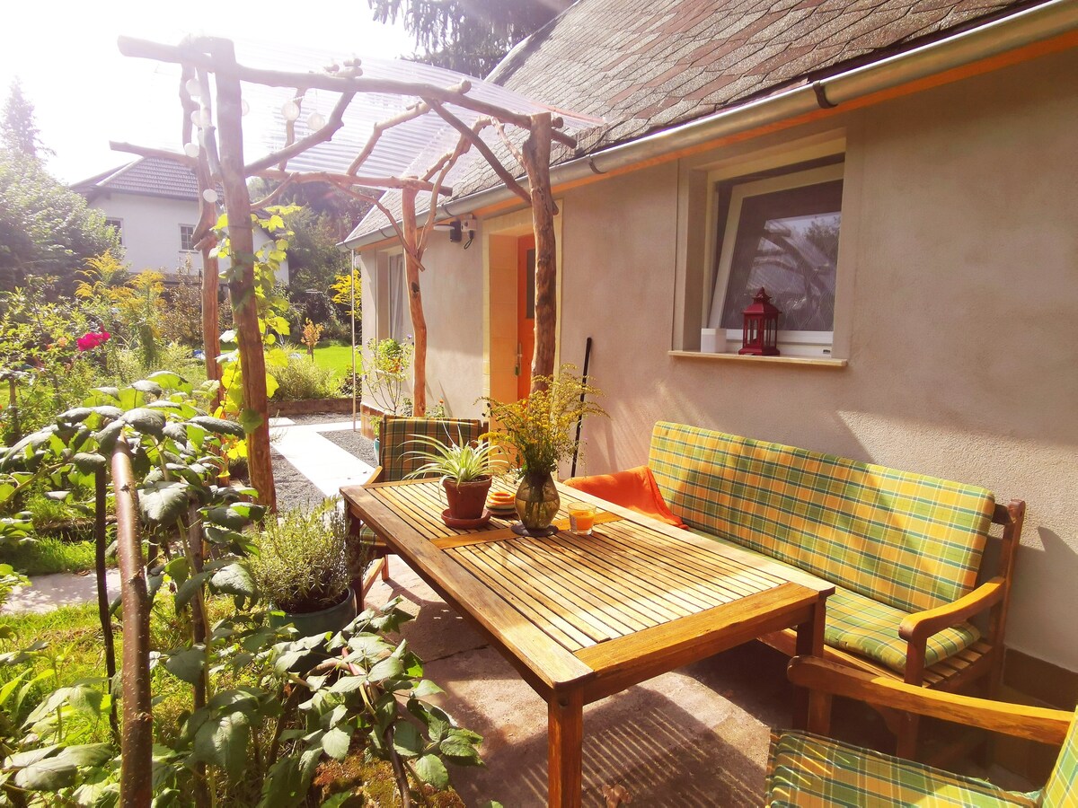 An outdoor seating area features a wooden table surrounded by cushioned chairs, complemented by potted plants. A trellis overhead supports climbing plants, and colorful garden blooms are visible nearby. The peaceful setting invites relaxation and enjoyment of nature.
