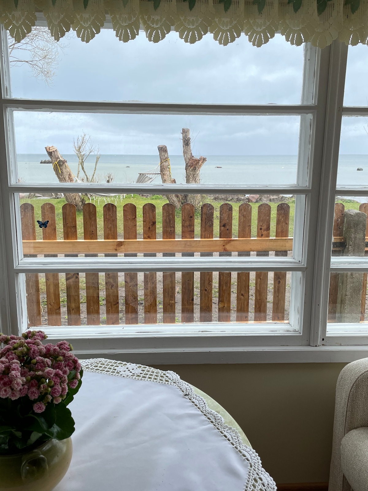 A view of the serene outdoor area is captured through a window. A wooden fence separates the property from the beach, which is visible in the distance. Green grass is seen in the foreground, complemented by a small bouquet of flowers on the table inside.