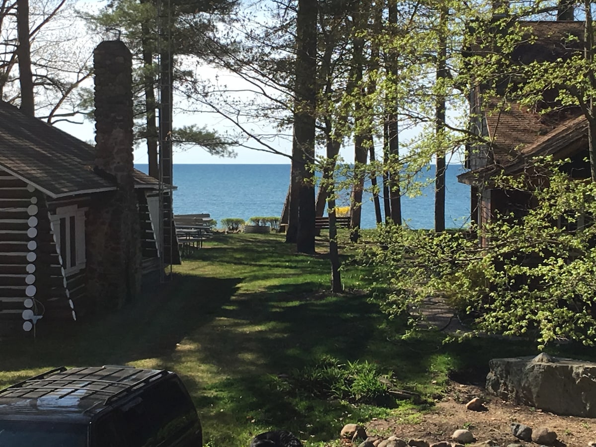 A serene pathway lined with trees leads toward a clear view of Lake Huron, with outdoor seating visible on the shore. Rustic cottages are framed by greenery, creating a peaceful atmosphere in this lakeside setting.