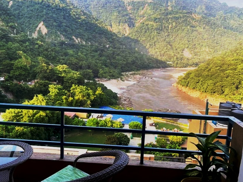 A scenic view from a balcony overlooks the Ganga river and lush green mountains. The landscape features a pool and garden area below, with the river winding through the valley. Comfortable seating is positioned on the balcony, inviting relaxation amidst natural beauty.
