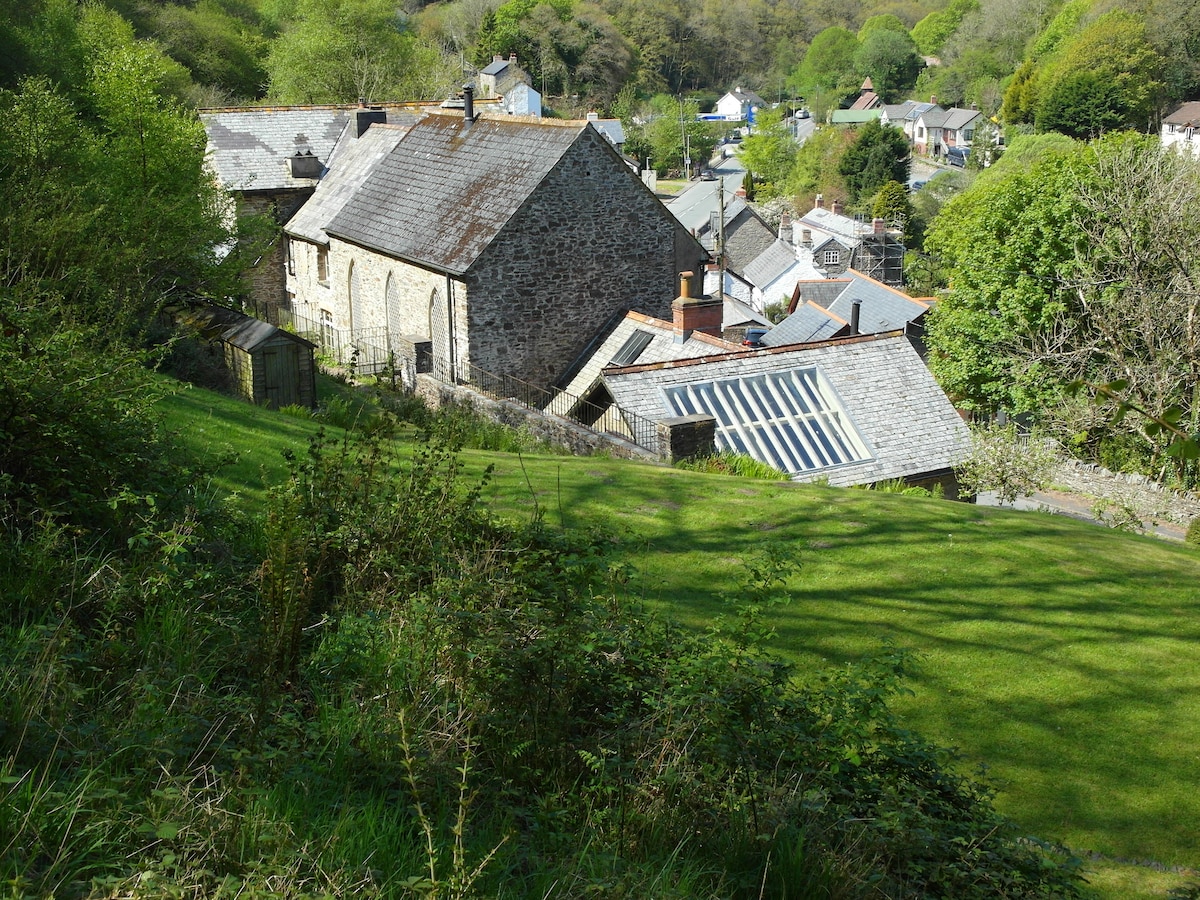 The image captures a hillside view of the Barbrook schoolroom and surrounding buildings, set in a lush green landscape. The diverse architecture of the structures, with sloping roofs and large windows, is framed by trees and open fields, creating a peaceful and natural setting.