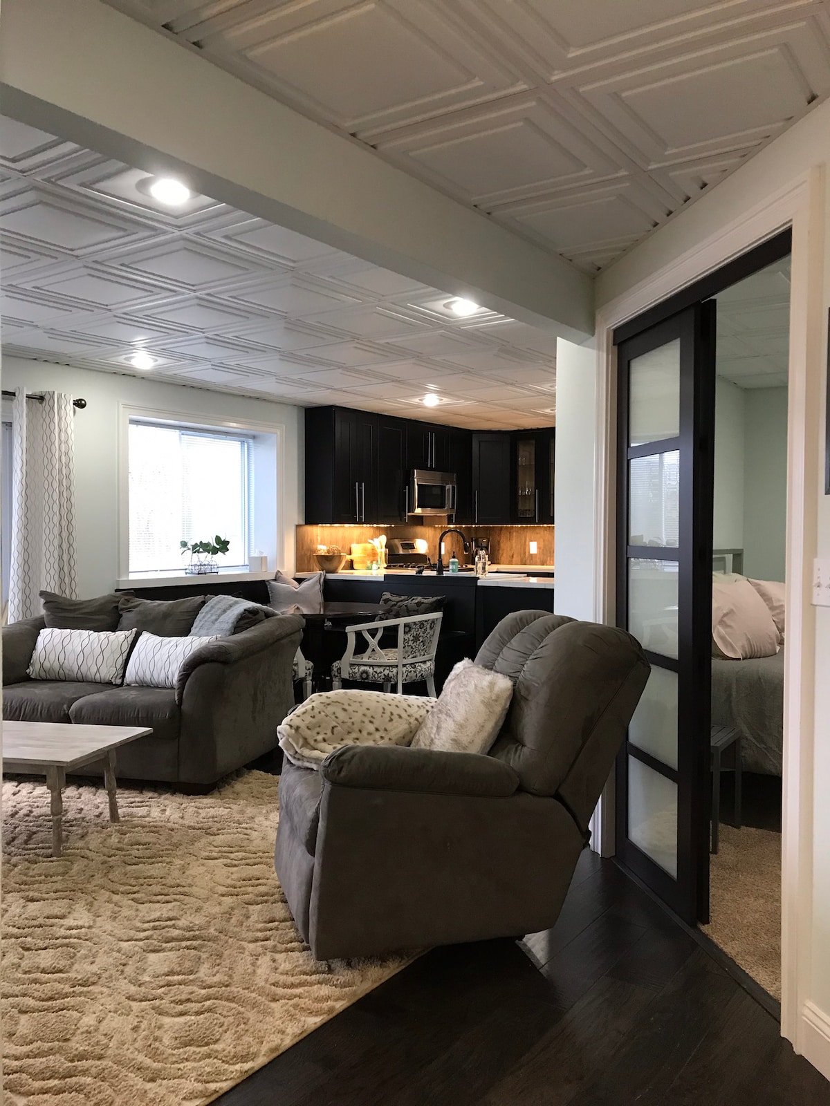 A spacious living area features comfortable seating including a gray sofa and an armchair, with a soft area rug underfoot. The kitchen is visible in the background, furnished with modern appliances and dark cabinetry. Natural light filters in through a large window.