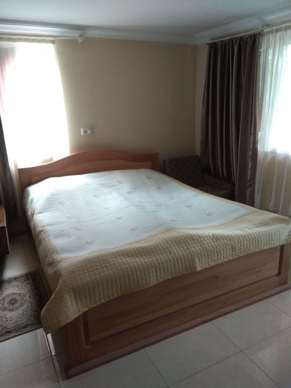 A cozy sleeping area features a spacious bed covered with a light quilt. A textured armchair is positioned nearby, and natural light enters through two windows adorned with sheer curtains.