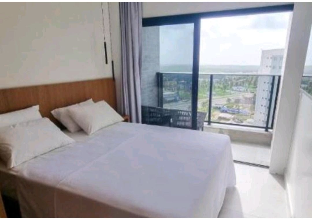 A bright bedroom features a spacious bed with white linens and two pillows. Large glass doors open to a balcony that offers views of the surrounding area. Natural light enhances the airy feel of the room, complemented by neutral-colored curtains.