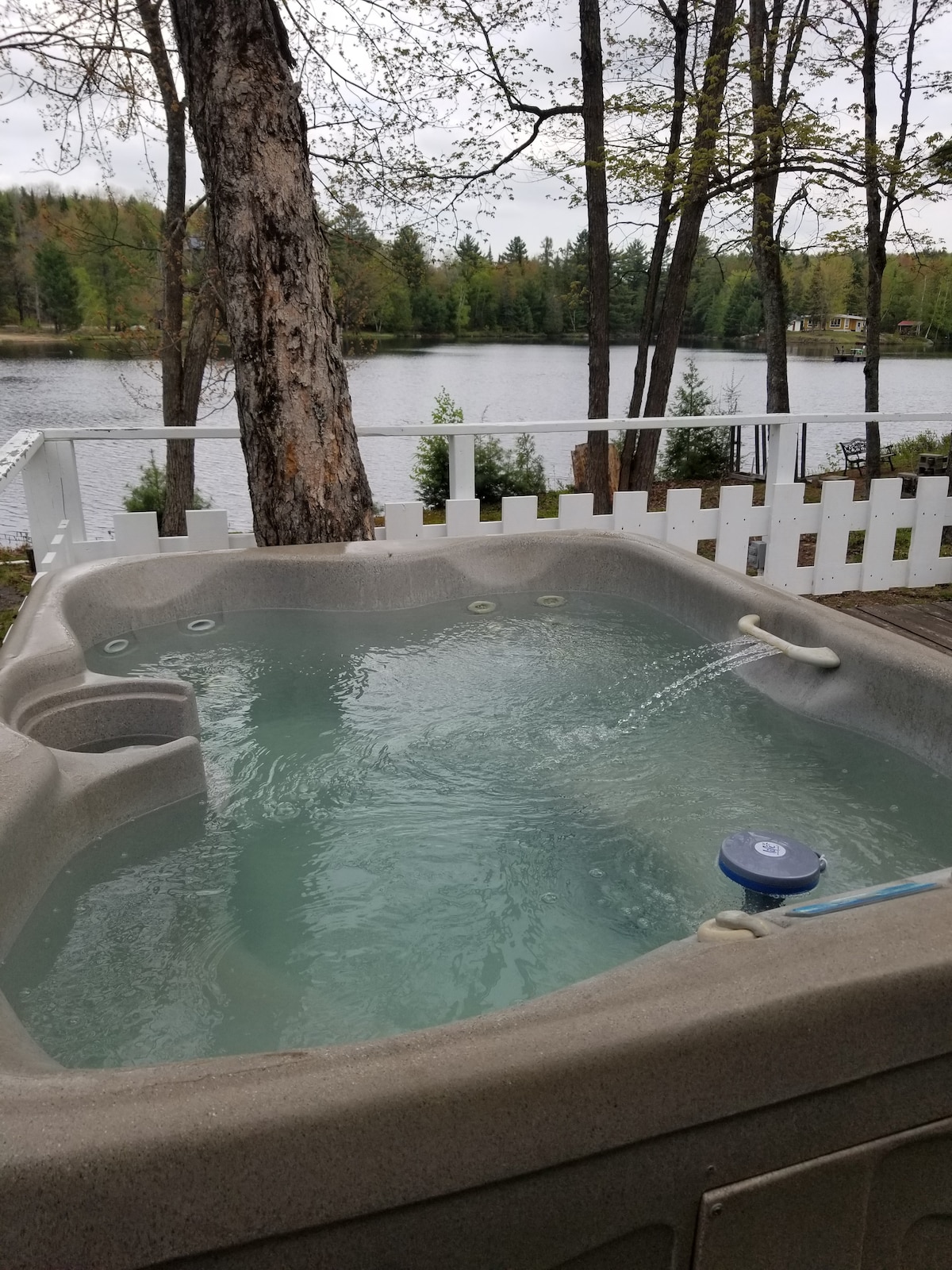 A private hot tub is situated on a wooden deck, surrounded by trees. The water appears clear and inviting, with gentle jets creating ripples. In the background, a peaceful lake reflects the greenery, with fencing providing a sense of privacy.