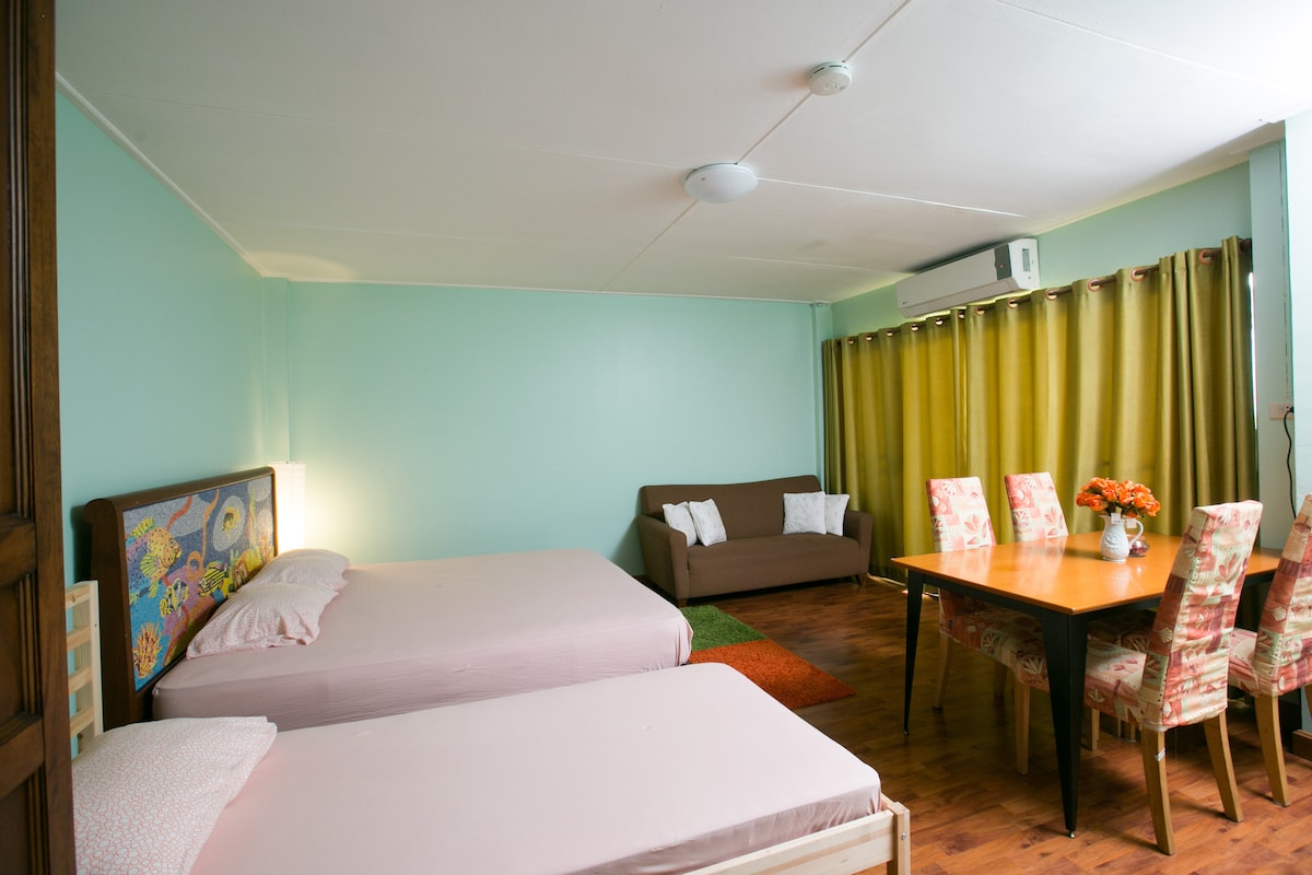 The room features two spacious beds with light bedding, complemented by a cozy brown sofa. A dining area with a wooden table and six patterned chairs is present. Natural light enters through large windows framed by floor-length curtains.