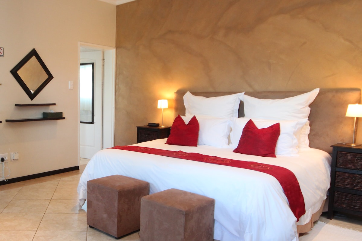 A spacious room features a king-size bed adorned with crisp white linens and decorative pillows in shades of red. Two ottomans are placed at the foot of the bed. Soft lighting is provided by lamps on each side, and a wall-mounted mirror adds depth to the space.