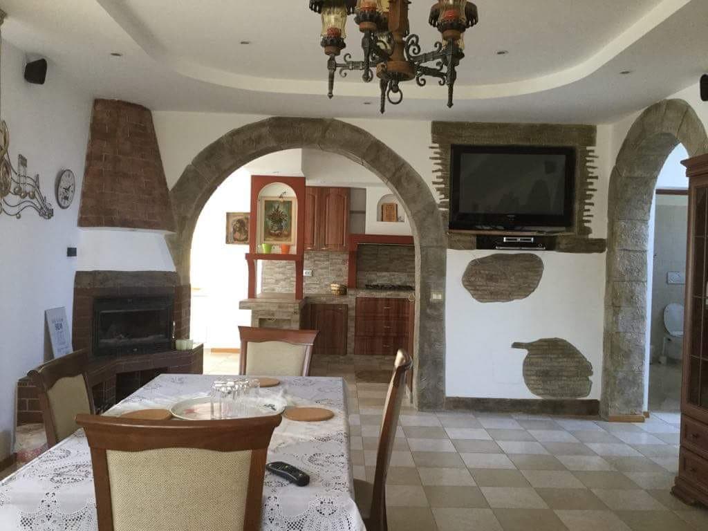 An open dining space features a large table with decorative settings, surrounded by upholstered chairs. Arched stone details frame the entrance to the kitchen area, where wooden cabinets and a flat-screen TV are visible. A cozy fireplace adds character to the room.