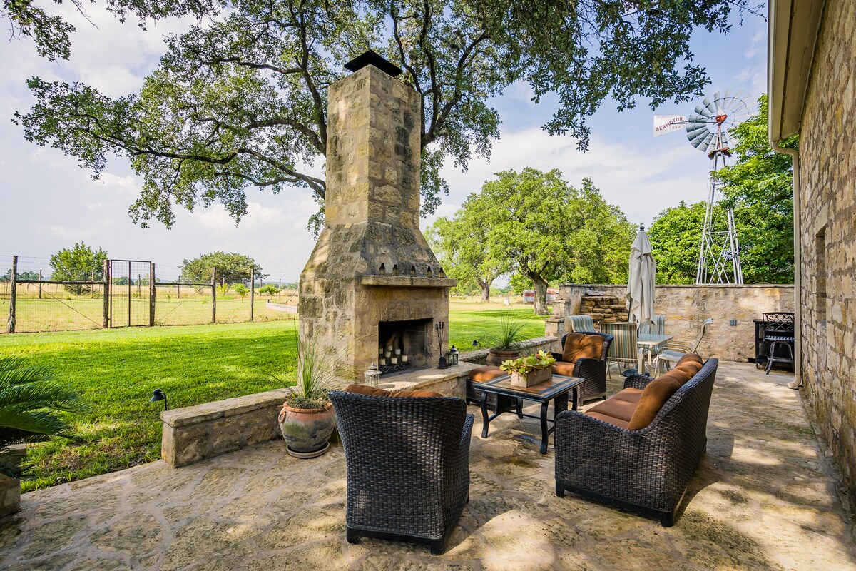 An outdoor patio area features a stone fireplace surrounded by comfortable seating. Lush green grass extends into the distance, where a rustic windmill and an enclosed area can be seen. Sunlight filters through the trees, creating a serene ambiance.