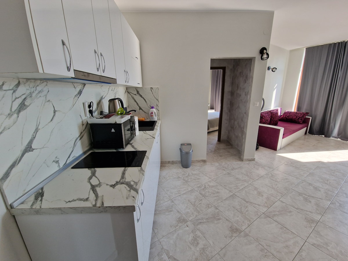 A modern kitchen with a marble countertop is visible, featuring a kettle and a compact cooktop. The room flows into a living area, distinguished by large windows that allow natural light. A glimpse of a bedroom can be seen through the doorway, along with a cozy seating area.