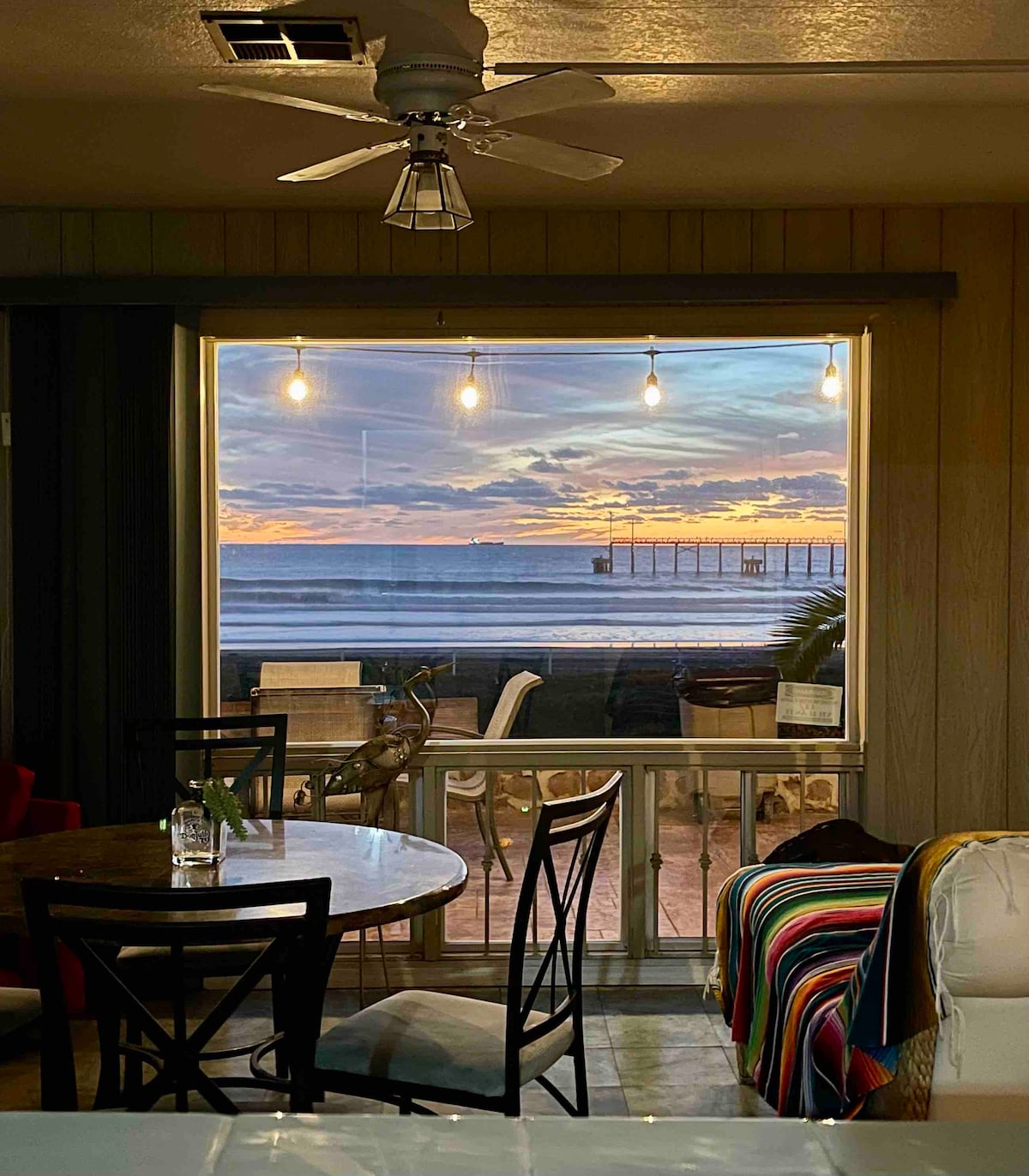 A dining area is visible, featuring a round table surrounded by black chairs. Natural light fills the space through a large window showcasing a view of the ocean and a pier at sunset. String lights add a cozy touch to the ambiance.