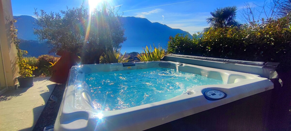 A private jacuzzi spa is situated in a well-maintained garden, surrounded by lush greenery. The sun shines brightly, reflecting off the water. Stunning mountain views are visible in the background, contributing to a serene outdoor setting.