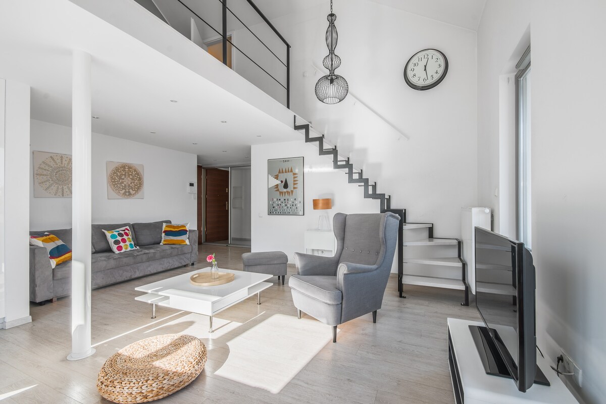 A light-filled living space is presented with neutral walls and wooden flooring. Comfortable seating is arranged around a coffee table, with a wall-mounted television nearby. A staircase leads to an upper level. Decorative elements include a modern clock and artwork, adding character to the room.