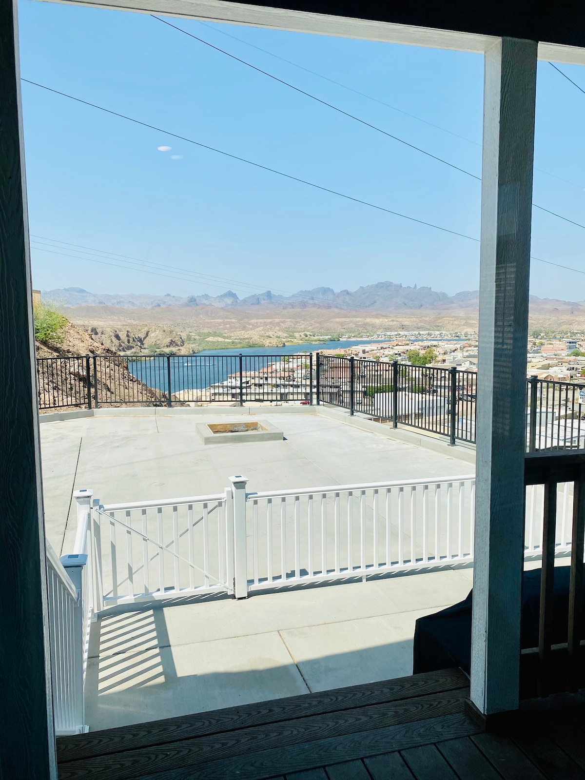 A view from inside the home features a spacious patio overlooking the Colorado River. The surrounding landscape includes mountains and open scenery. A white fence encloses the area, while a fire pit is centered on the concrete patio, ready for gatherings.