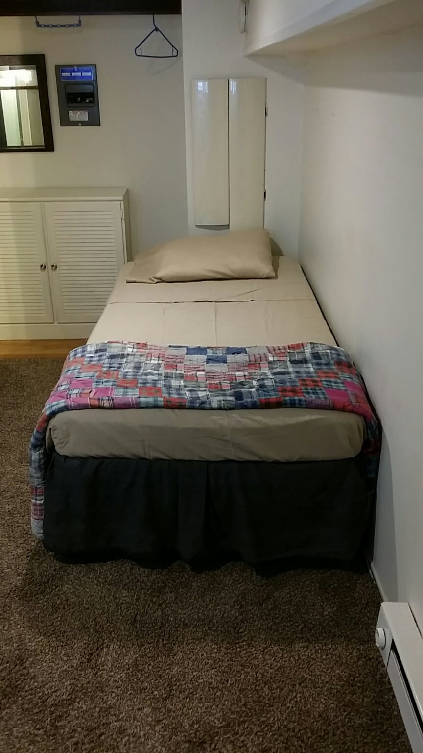 A small bedroom area features a single bed dressed with a colorful quilt. A simple bedside table and wall-mounted mirror are visible, providing functionality. Soft lighting enhances the room's peaceful ambiance, while the carpeted floor adds warmth to the space.