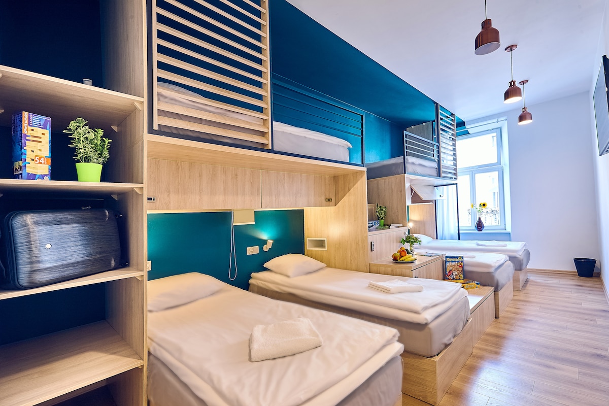 A spacious room features six comfortable beds with safe surroundings and personal storage. Light wood cabinetry complements the vibrant blue walls. Two large windows provide natural light, while modern lighting fixtures add to the ambiance. A flat-screen TV is mounted on the opposite wall.