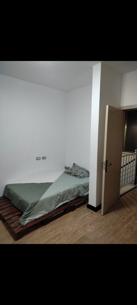A simple bedroom is presented with a bed on a wooden platform, featuring a light green blanket and decorative pillows. White walls enhance the space, while a door leads to another area, allowing natural light to enter.