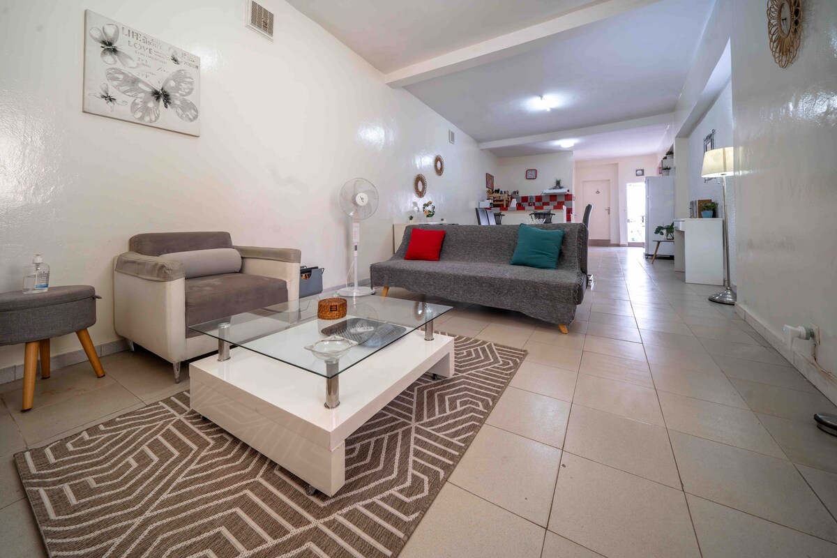 The living area features a spacious layout with a gray sofa and a comfortable armchair, complemented by colorful throw pillows. A glass coffee table sits on a patterned rug, while a fan provides air circulation. In the background, the kitchenette and dining area are visible.
