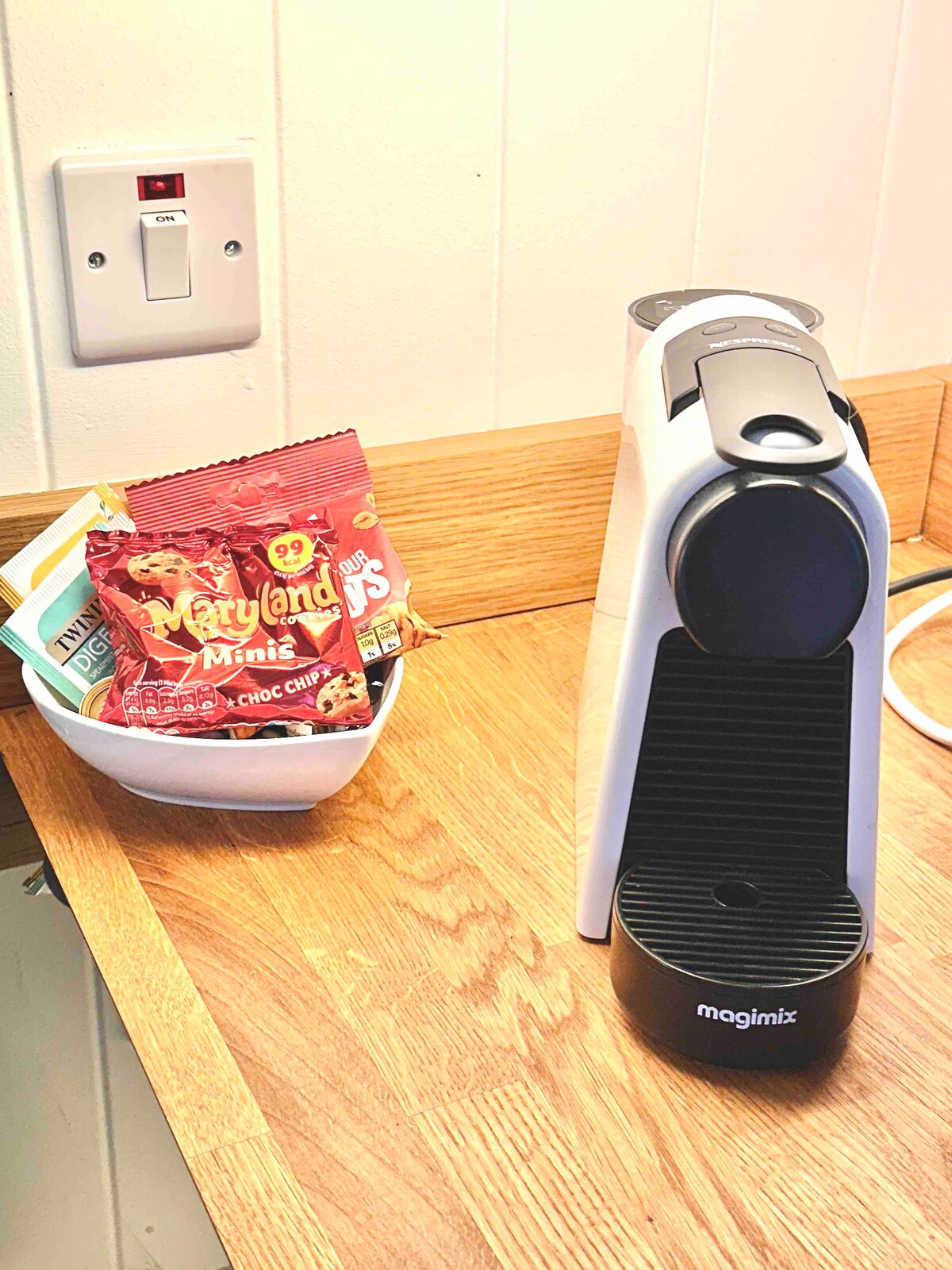 A modern coffee machine with a sleek design is positioned on a wooden countertop. Next to it, a white bowl contains a selection of chocolate candies, providing a cozy touch to the kitchen area.