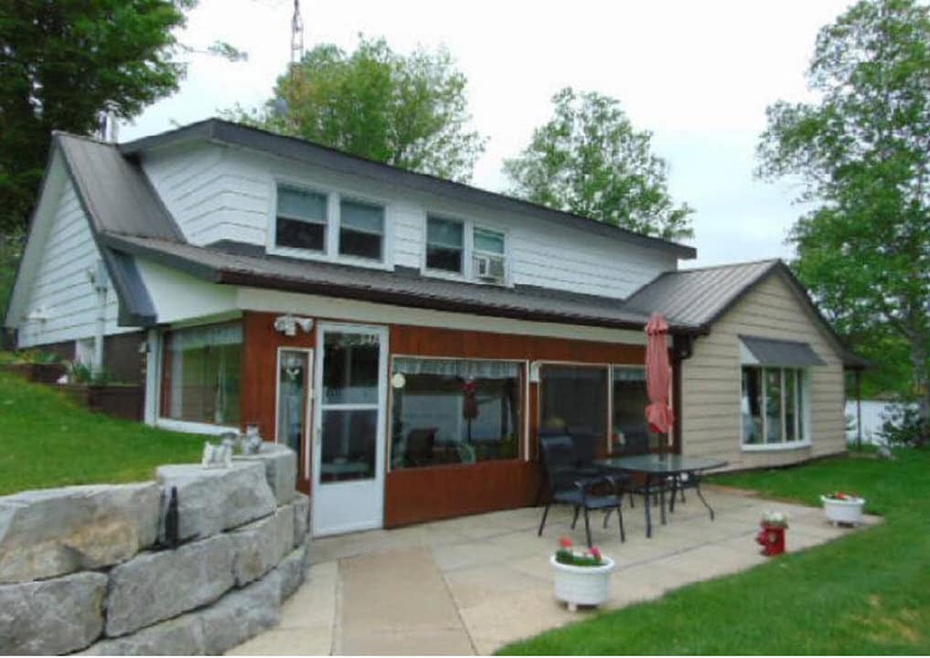 The exterior of the house features a blend of white and beige siding with a dark metal roof. A stone pathway leads to a patio area furnished with a table and chairs, complemented by potted flowers. Lush greenery surrounds the property, enhancing the natural setting.