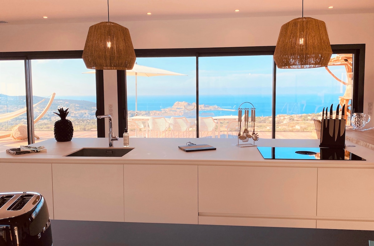 A modern kitchen space features a large island with a sink and cooktop, accented by two woven pendant lights. Large windows frame a stunning ocean view, allowing natural light to illuminate the interior, while kitchen utensils and a stylish fruit centerpiece are visible on the counter.