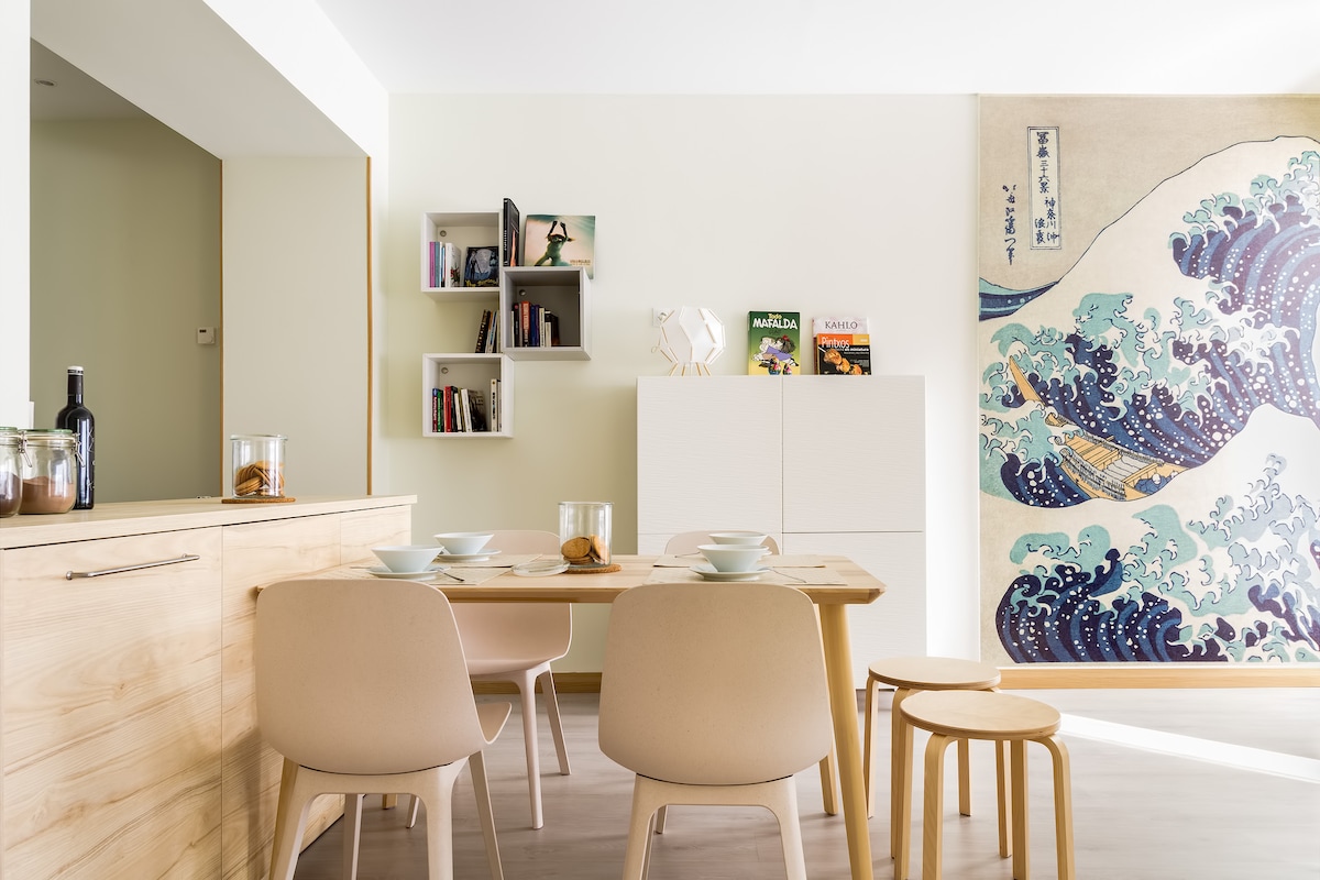 A dining area is presented with a wooden table set for breakfast, surrounded by light-colored chairs. A large, vibrant wall art depicts waves, contributing to the space's atmosphere. Shelves are displayed with books and decorative items, emphasizing a cozy yet modern aesthetic.