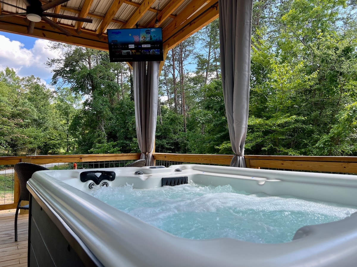 The hot tub is situated on a wooden deck, featuring jets and bubbling water. A flat-screen TV is mounted above, with tall curtains providing privacy. Lush greenery is visible in the background, adding to the serene outdoor setting.