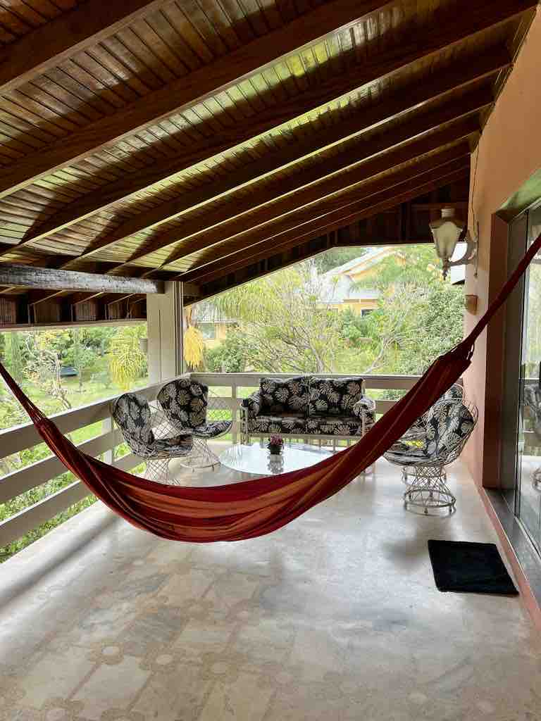 A spacious covered balcony offers a relaxed seating area, featuring a floral patterned couch, two wicker chairs, and a glass table. A vibrant red hammock hangs nearby, with lush greenery visible in the background, providing a serene outdoor atmosphere.