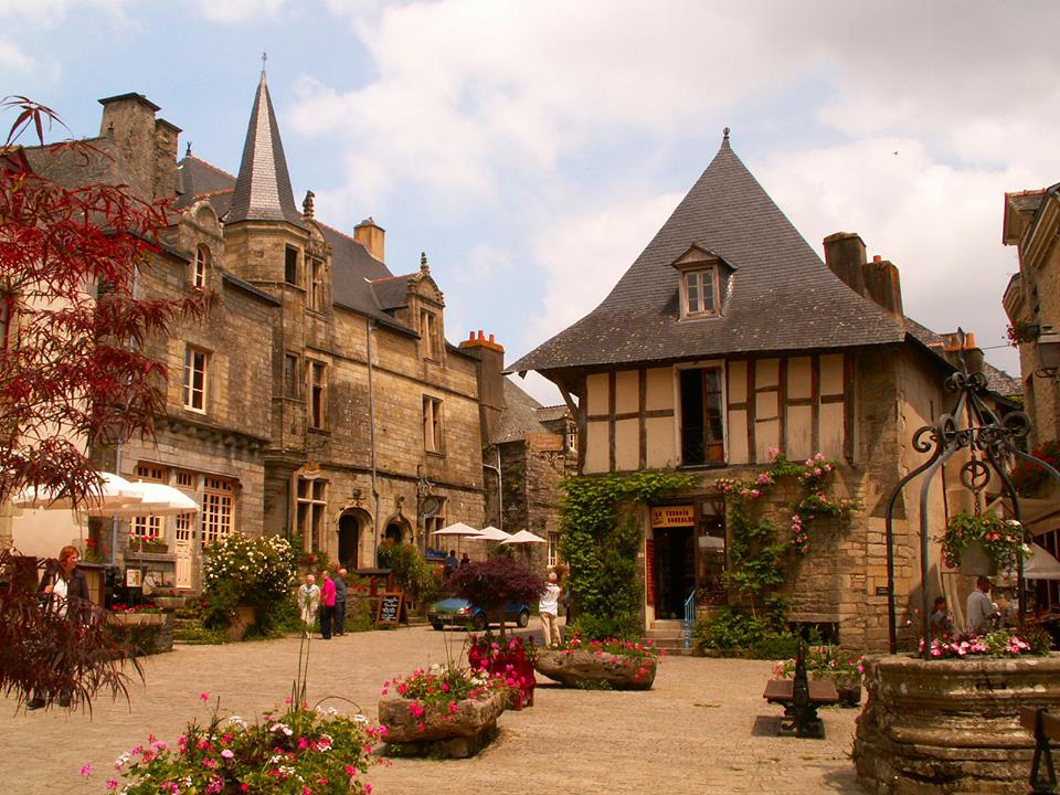 A charming village square is depicted, surrounded by historic stone buildings with varied architectural styles. Colorful flowers border the pathways, and café tables are set up outside. Patrons enjoy the serene atmosphere under a partly cloudy sky.