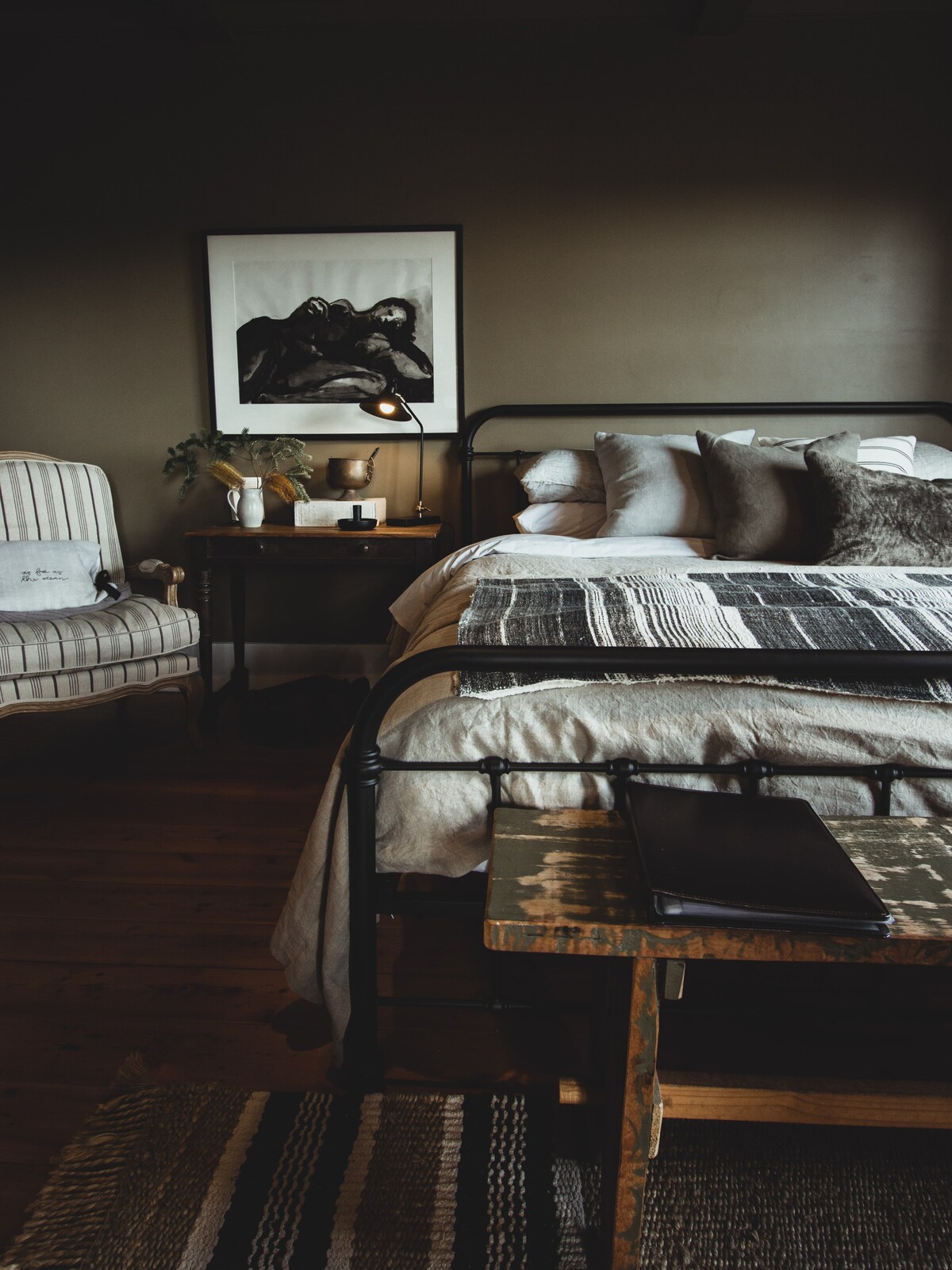 A king cast iron bed covered with soft linens is positioned next to a wooden side table featuring a stylish lamp. An inviting armchair with stripes complements the setting, while an artistic black-and-white print hangs above the bed, enhancing the room's elegant tones.