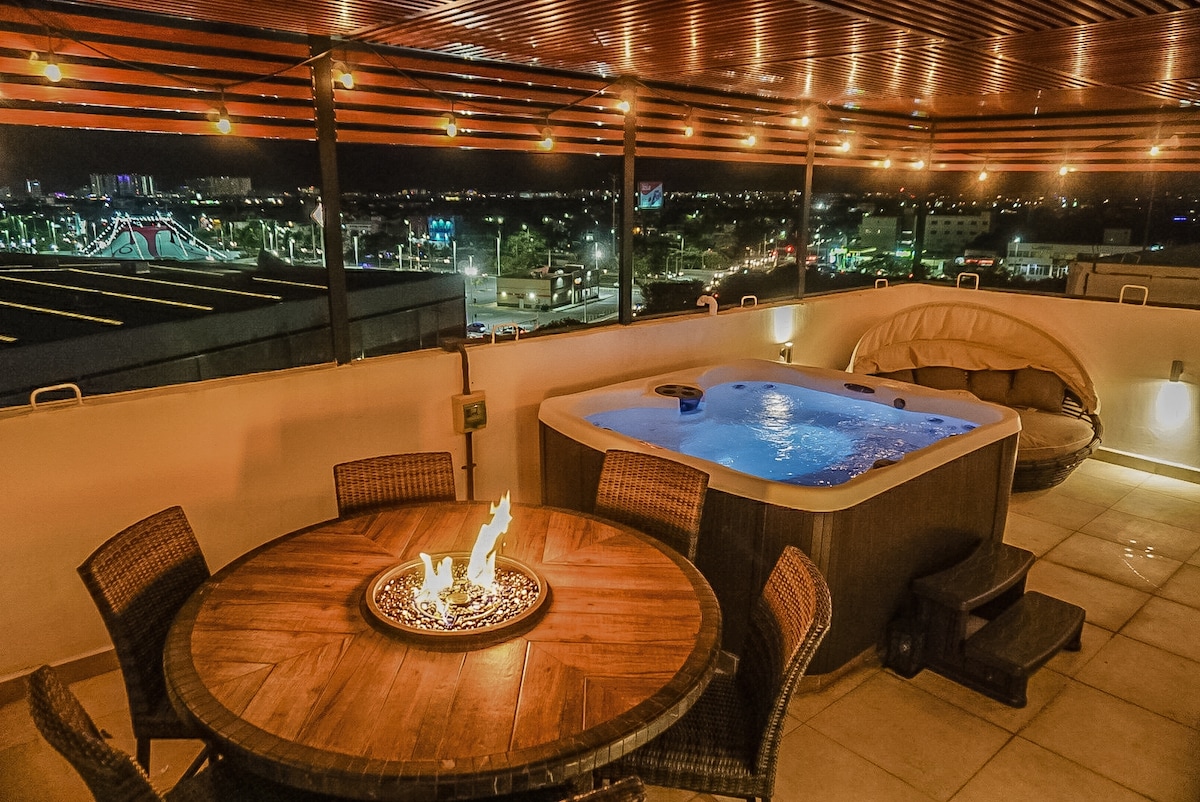 A private rooftop area features a circular wooden table surrounded by woven chairs, a glowing fire pit at its center, and a hot tub with bubbling water. String lights provide soft illumination, creating an inviting atmosphere with views of the city skyline.