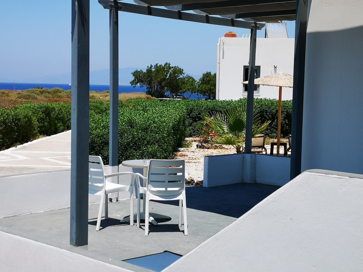 A shaded outdoor seating area features two white chairs and a round table, surrounded by lush greenery and a view of the distant sea. A sun umbrella provides additional shade, enhancing the space for relaxation.