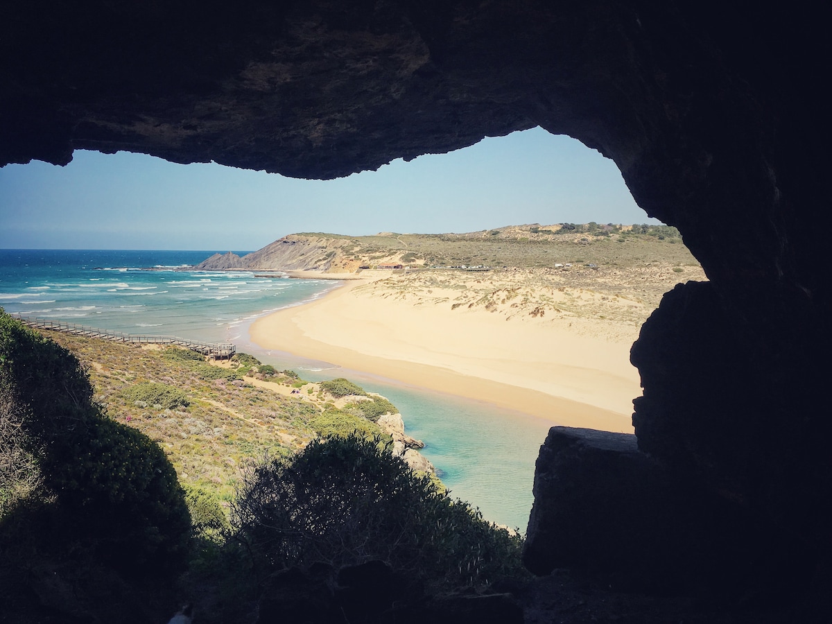 A natural rock formation frames a view of a serene beach and ocean. Soft waves gently lap at a sandy shore, while verdant vegetation surrounds the scene. The expansive coastline stretches into the background, highlighting the beauty of the surrounding landscape.
