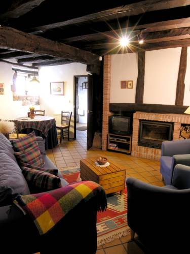 A warm living area features dark wooden beams overhead, with a blue sofa adorned with a colorful throw blanket. A rustic coffee table sits on a patterned rug, alongside two blue armchairs. The space is illuminated by a soft overhead light, creating a cozy atmosphere.