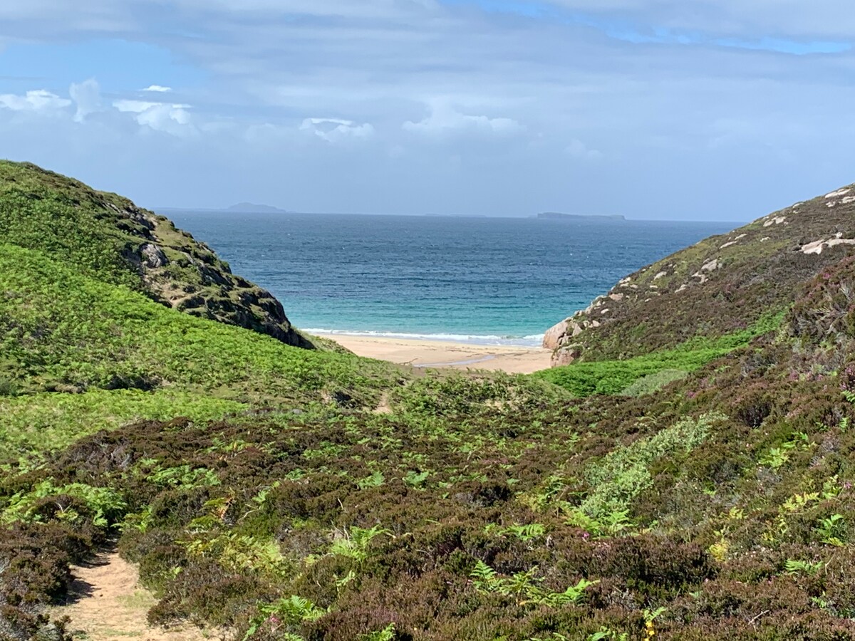 A scenic view captures the approach to a sandy beach flanked by lush green hills. The calm sea stretches into the distance, with waves gently lapping at the shore. A clear sky is visible overhead, enhancing the tranquil coastal landscape.