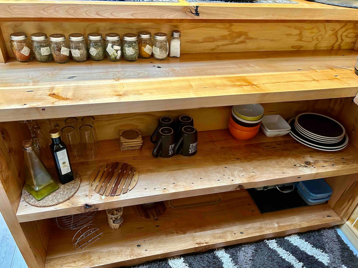 A wooden storage unit displays various jars containing spices on the top shelf. The lower shelves hold glassware, brown mugs, colorful plates, and additional containers. A small bottle of olive oil and kitchen utensils are also present, enhancing the functionality of the space.