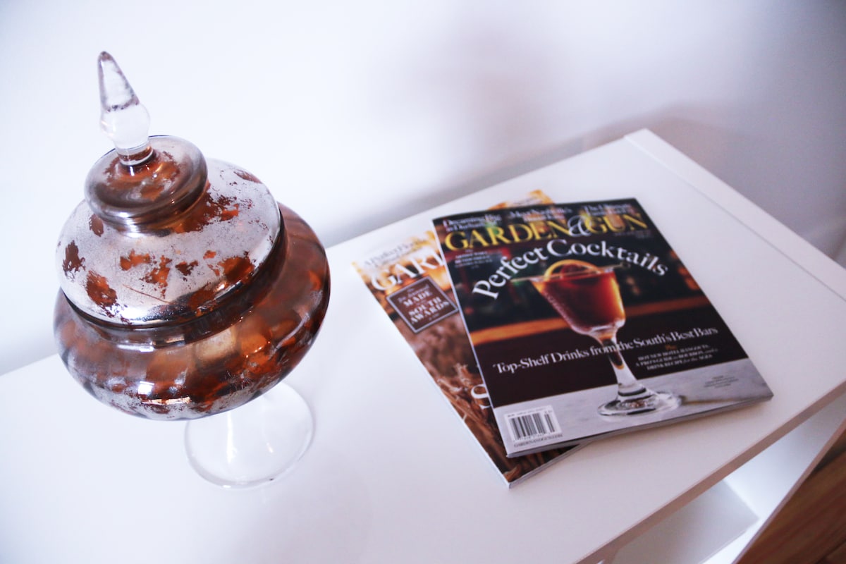 A clear glass table is adorned with a decorative glass jar featuring an ornate lid. Nearby, a stack of magazines showcases a cocktail-themed publication, highlighting drink recipes and ideas for entertaining. The overall arrangement conveys a sense of casual sophistication.