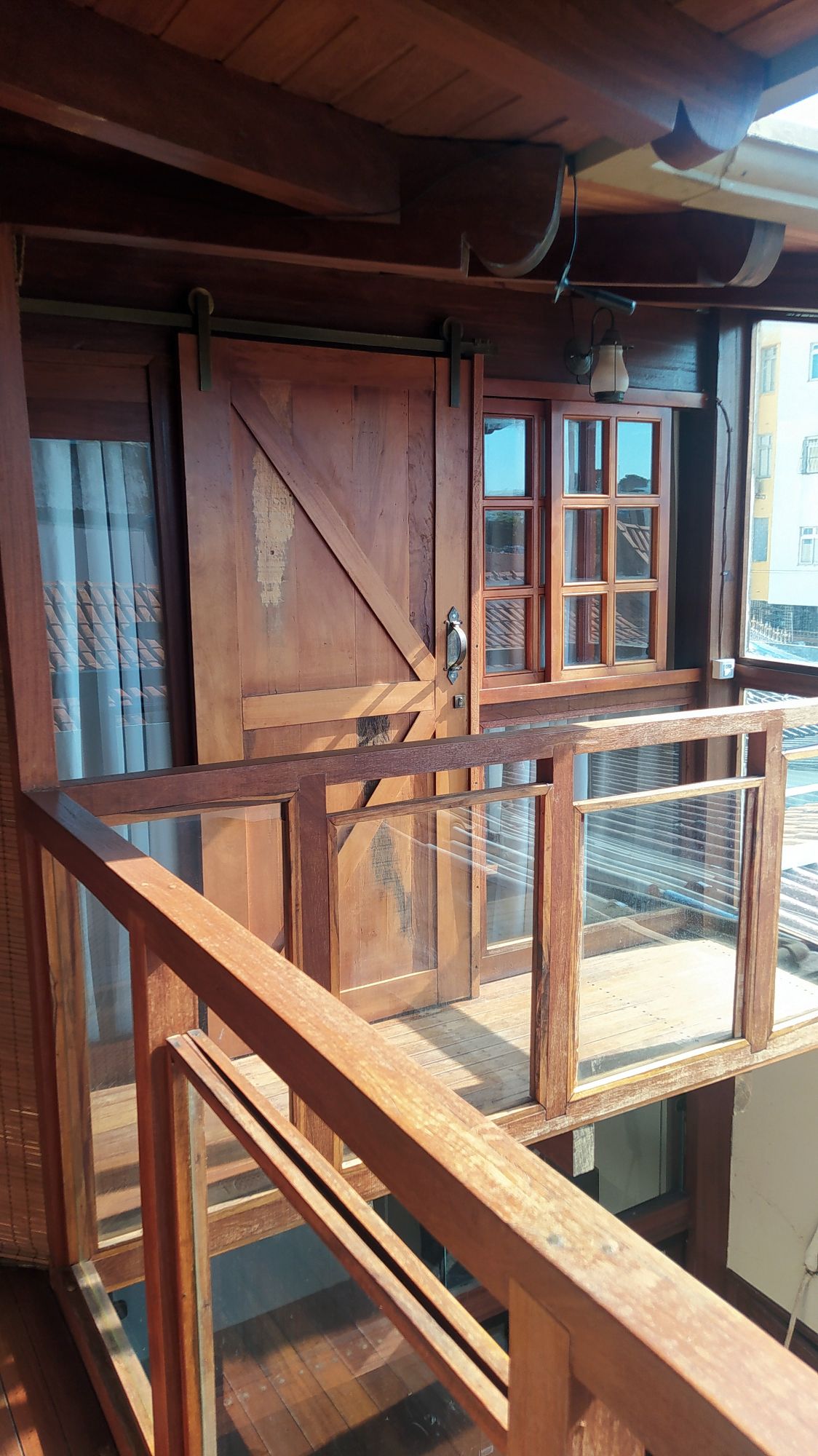 A wooden balcony features transparent railings and sliding doors, providing access to interior spaces. Natural light illuminates the area, highlighting the rich wood tones. Nearby, a traditional window adds charm, contributing to a cozy atmosphere.