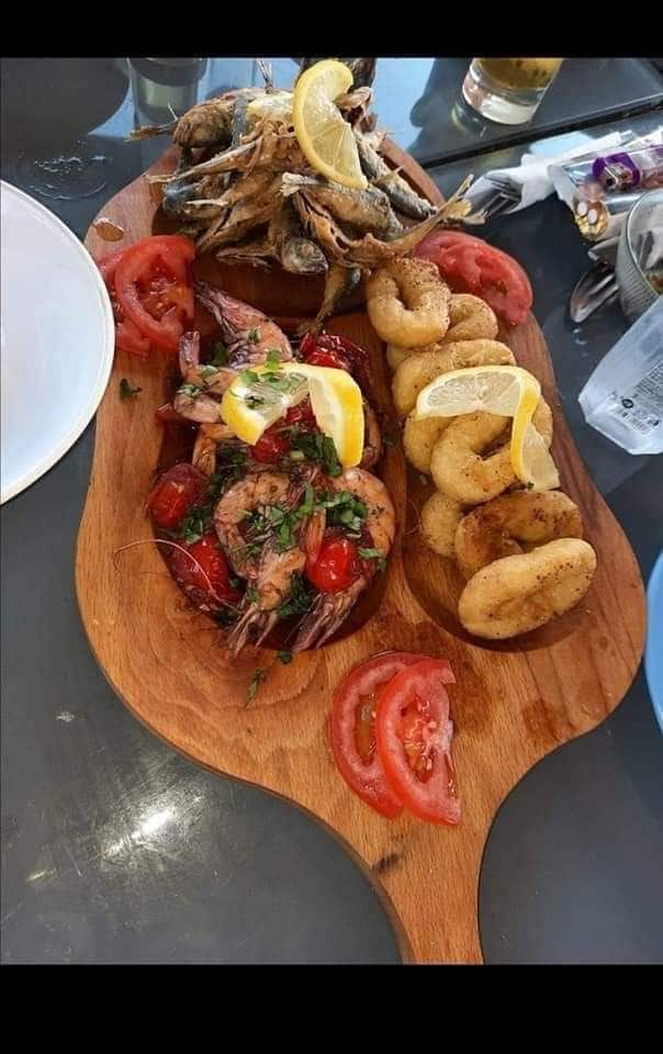 A wooden platter displays a variety of seafood, including shrimp garnished with tomatoes and herbs, alongside golden fried items. Lemon wedges are placed atop the seafood, with sliced tomatoes arranged around the dish. The setting hints at a casual dining experience.