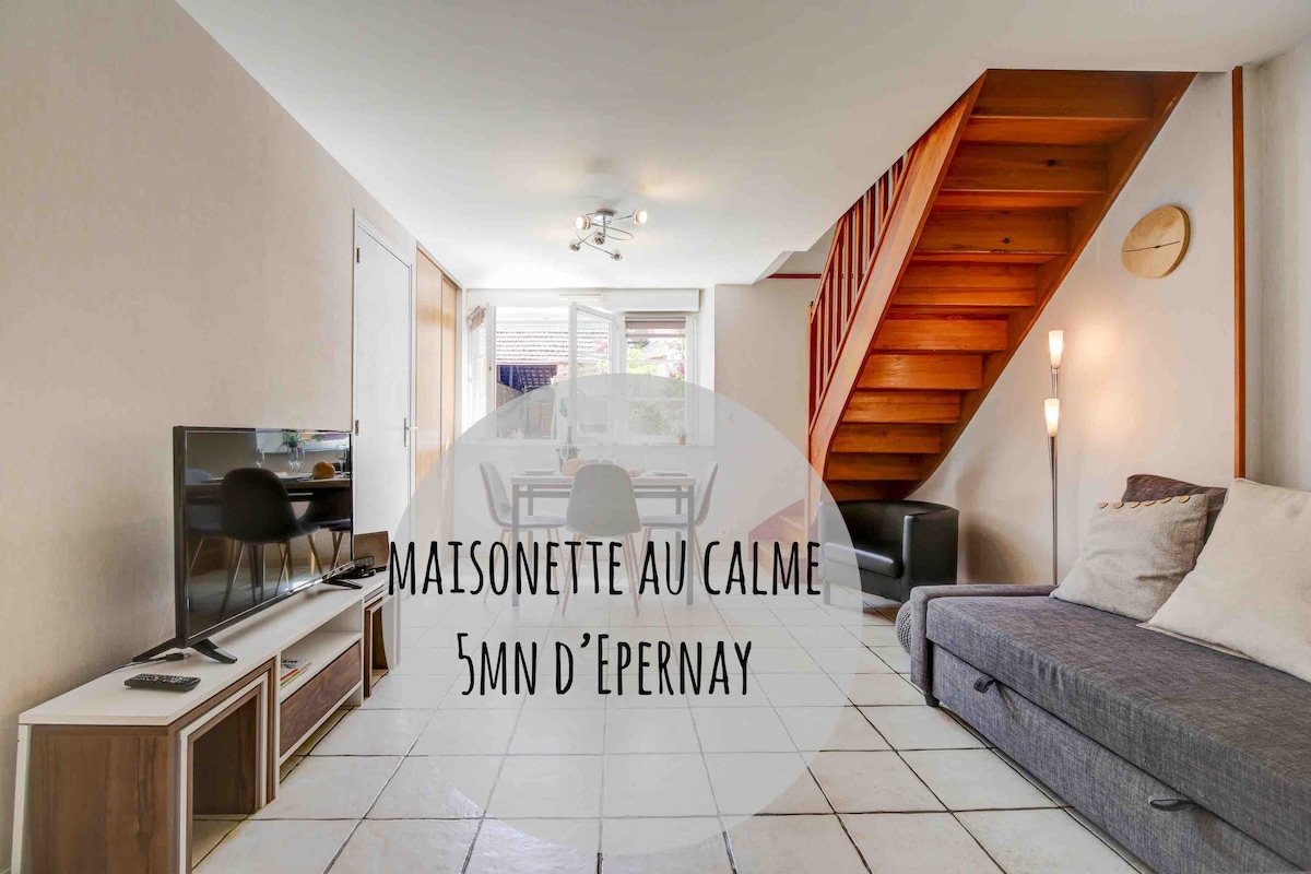 A cozy living space is showcased, featuring a sectional sofa and a television on a light wood media console. Large windows allow natural light to fill the room. A staircase with wooden steps leads to the upper floor, complementing the neutral tones of the decor.