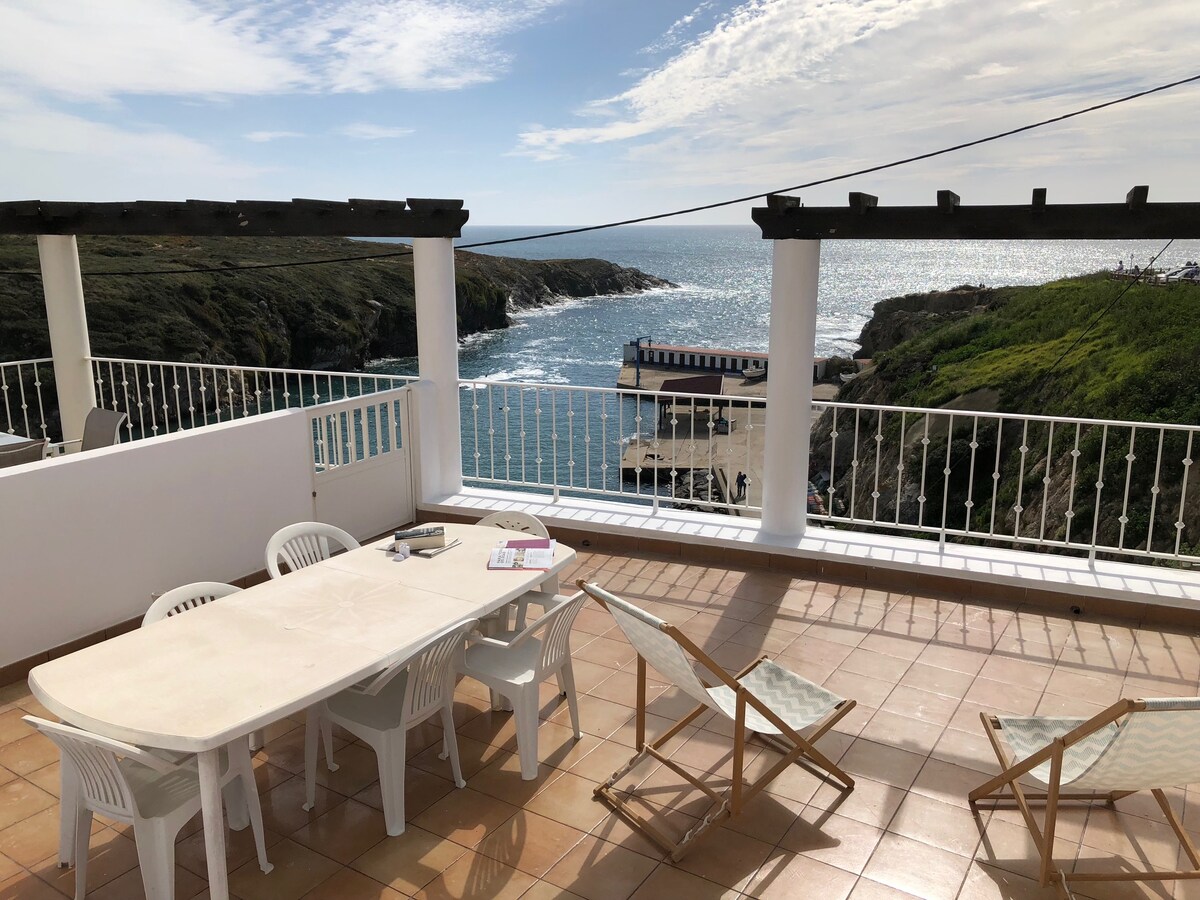 A spacious terrace features a large dining table surrounded by several white chairs. Sun loungers are placed nearby, providing a view of the blue sea and coastal landscape. The sky is partly cloudy, contributing to a bright and open atmosphere.