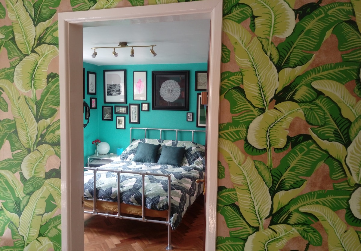 A bedroom is framed by a doorway, featuring vibrant green foliage wallpaper. A spacious bed, adorned with patterned bedding and coordinating pillows, is positioned against the wall. Surrounding the bed, framed art pieces add character to the room, complementing the playful decor.