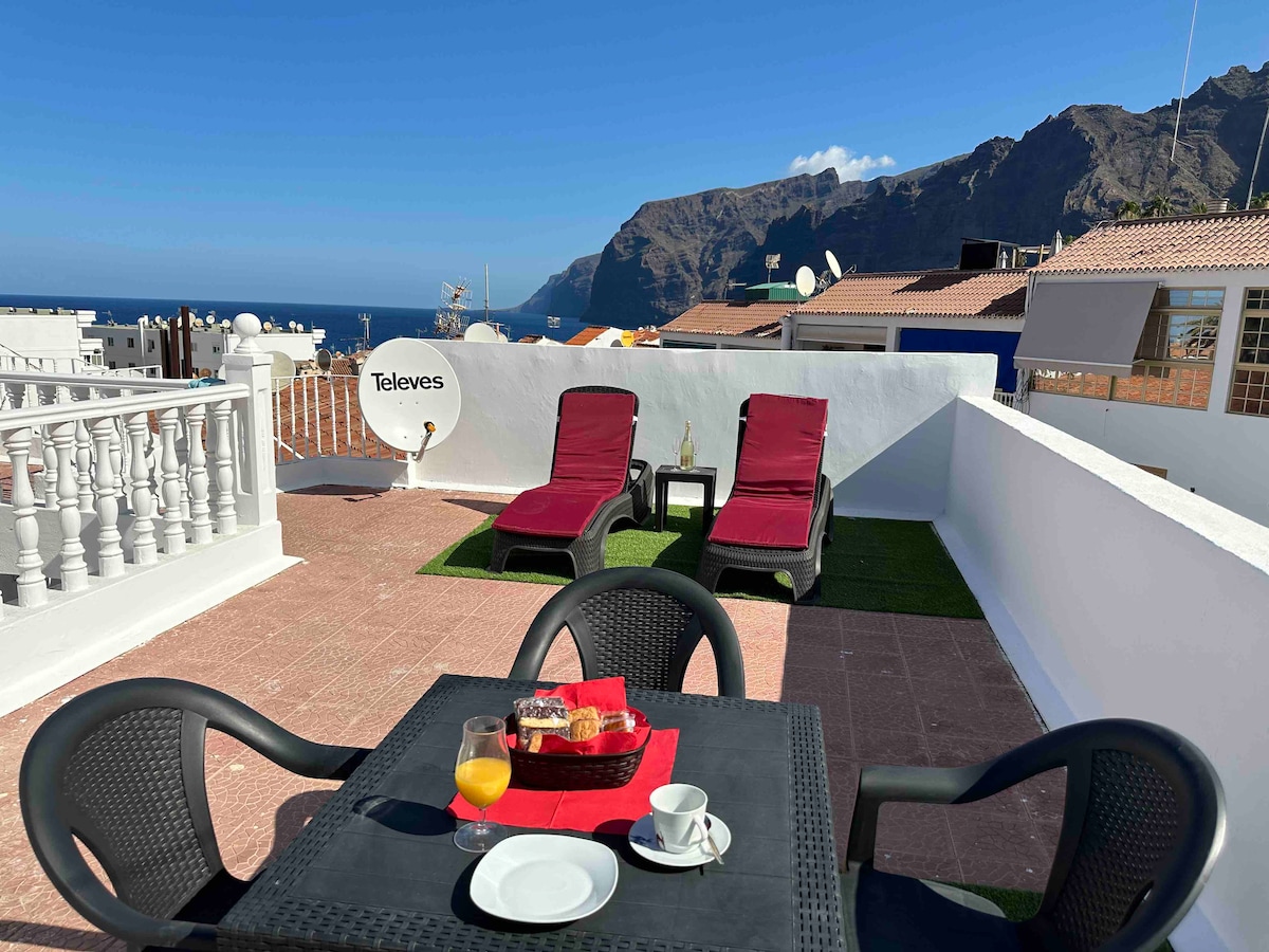 A rooftop terrace is presented with two lounge chairs and a small dining table. The table is set with a red cloth and includes a tray with pastries and a glass. Scenic views of mountains and the ocean are visible in the background.