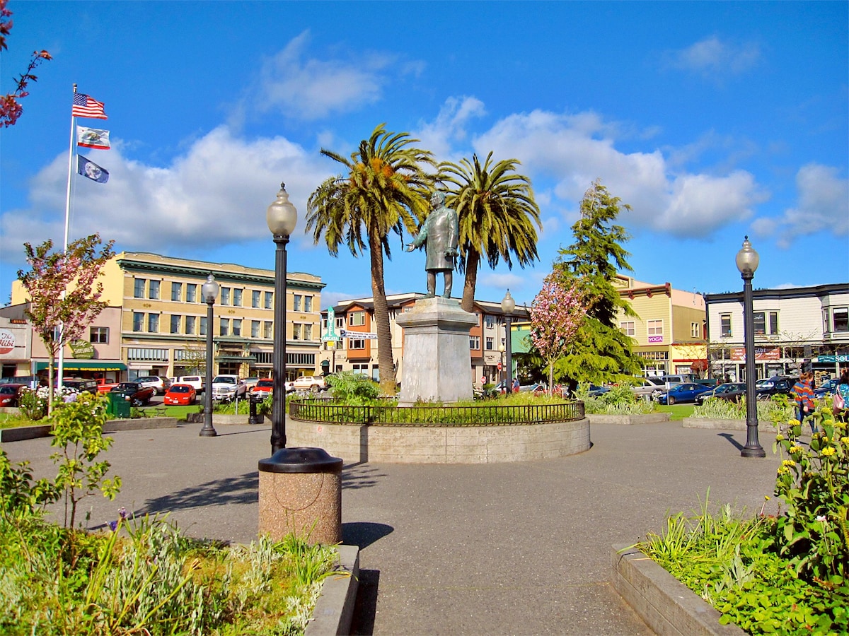 A vibrant public square features a statue surrounded by palm trees and blooming flowers. Buildings line the perimeter, showcasing various architectural styles. Flags flutter in the gentle breeze. The area is well-maintained, with benches and green spaces contributing to a welcoming atmosphere.