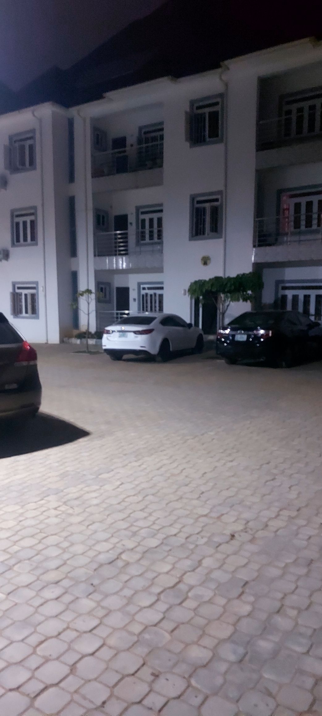 An illuminated parking area is visible, featuring several cars parked on a neatly paved surface. The white and black vehicles are positioned in front of a multi-story building, which is accented by soft outdoor lighting. The overall environment appears organized and well-maintained.