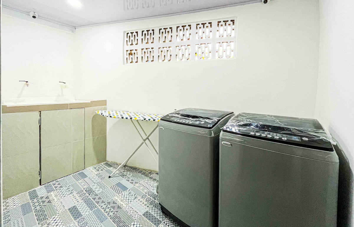 A functional laundry area showcases two modern washing machines positioned next to a countertop sink. An ironing board is neatly set up nearby, and patterned tiles cover the floor, adding a touch of design to the practical space.