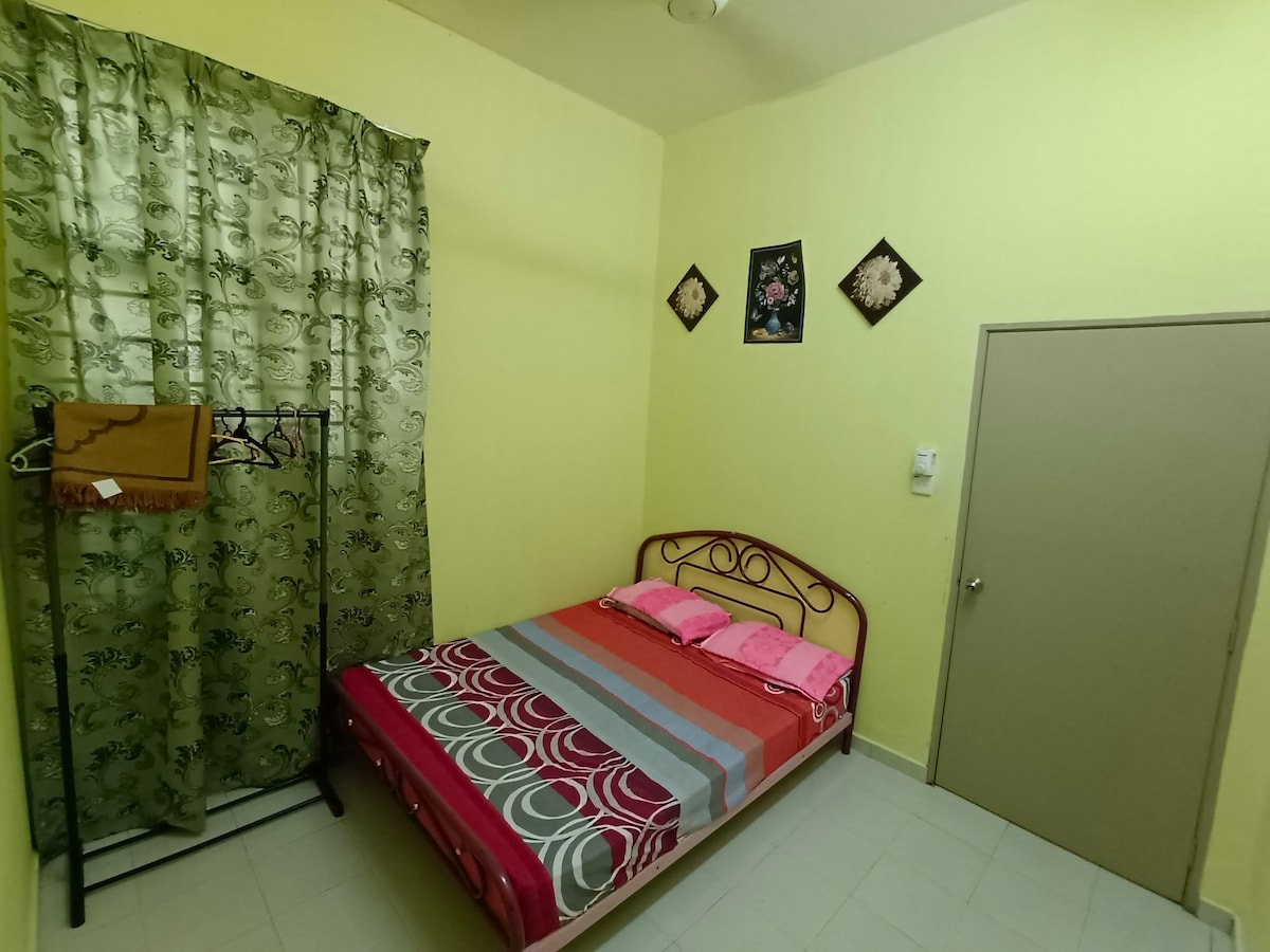 A bright guest room features a spacious bed adorned with colorful patterned bedding and pink pillows. Light green curtains frame a nearby window, while decorative wall hangings add character to the soft yellow walls. A clothing rack is positioned next to the bed for convenience.
