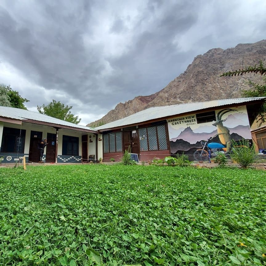 A guesthouse is situated against a backdrop of majestic mountains. The structure features a mural, large windows, and a landscaped lawn covered in lush greenery. A cloudy sky blankets the scene, adding depth to the overall view.