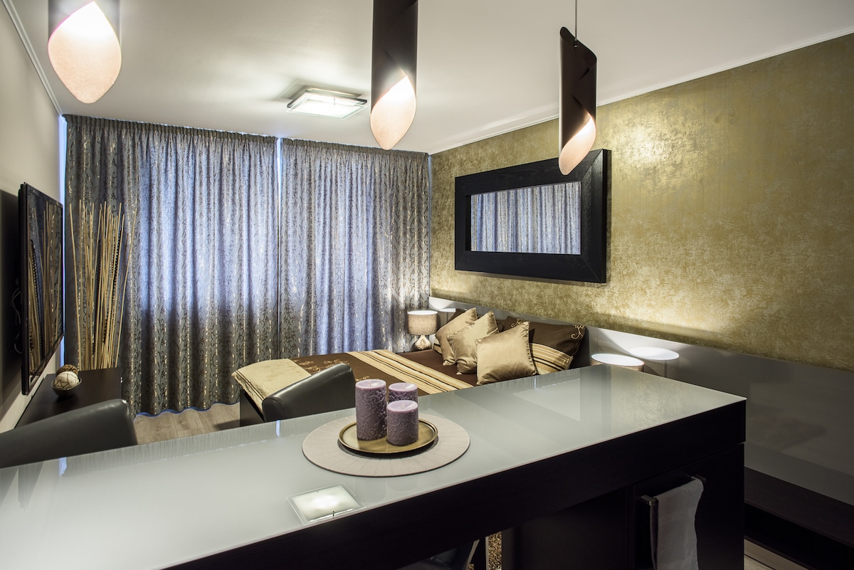 An open living area features a modern bar counter with decorative candles and a circular tray. Stylish lighting hangs above, while large curtains frame a cozy sleeping area with a textured wall and plush bedding.